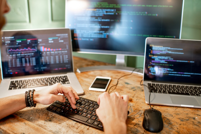Photo of laptop and computer screens and a man's hands typing computer programming code.