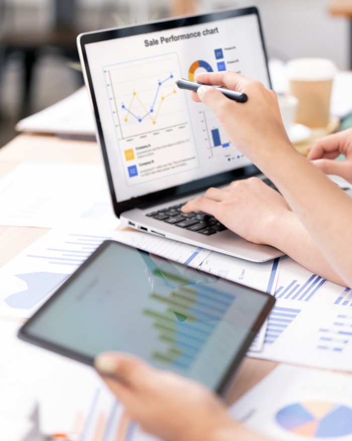 Photo of tablet and laptop and paper reports, the tablet has a graph on the screen and the laptop has graphs and charts.
