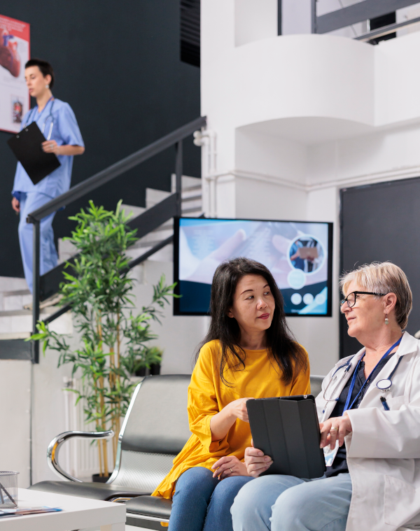 Photo of doctor and patient talking whilst sat down and medical staff on the stairs, in the background within a clinic.